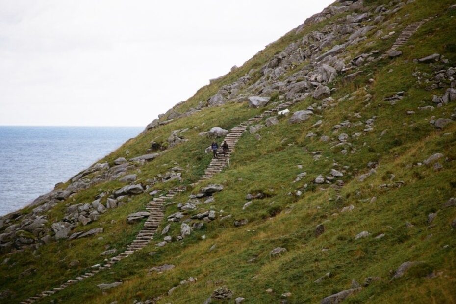 Two hikers climb stone steps on a steep grassy coastal hillside
